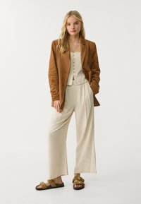 Brun mockajacka över en beige kort knappad topp, kombinerad med ljusbeige vida byxor och bruna sandaler med gulddetaljer.