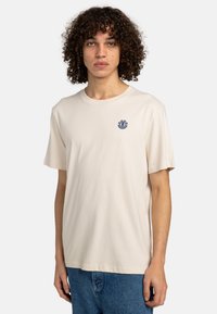 Beige Baumwoll-T-Shirt mit rundem Ausschnitt, kurzen Ärmeln und kleinem blauen Logo auf der linken Brust. Weiche Textur und lässige Passform.