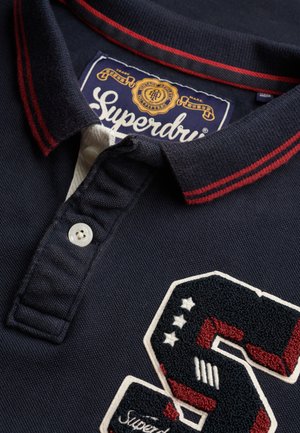 Polo bleu marine avec col rayé rouge, bouton blanc, patch texturé avec un "S" et des étoiles, et étiquette Superdry.