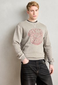 Grå hettegenser laget av mykt materiale, med et rødt sirkulært grafisk trykk som sier "TWENTY TWO SCOTCH & SODA," sammen med mørke jeans.