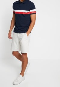 Hombre con polo azul marino a rayas blancas y rojas, pantalones cortos blancos y zapatillas blancas, de pie con una mano en el bolsillo.