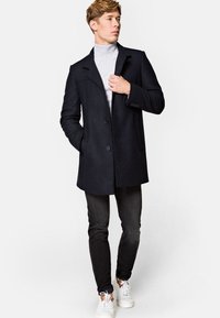 Płaszcz z wełny w ciemnym granacie z usztywnionym kołnierzem, z zamknięciem na pojedynczy guzik i kieszeniami po bokach, noszony na jasnoszarym golfie.