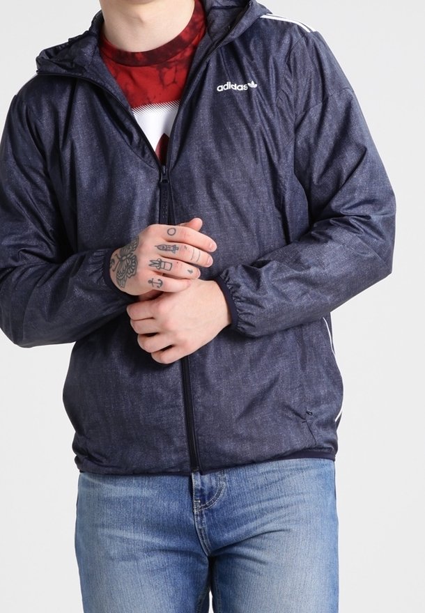Marinblå Adidas-jacka med texturerad yta, dragkedja fram och vita detaljer på axlarna. Bärs över en röd och vit mönstrad skjorta.