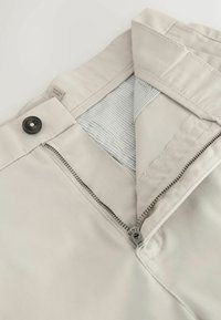Ceinture de pantalon beige défaites avec fermeture par bouton, fermeture éclair en métal et doublure intérieure rayée visible.