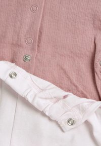 Světle růžová žebrovaná látka se zapínáním na patentky, překrytá bílou měkkou látkou s elastickým páskem. Viditelný detail knoflíku zvýrazňuje texturu švu.