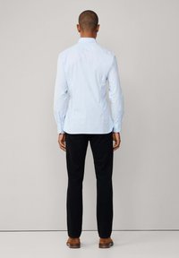 Muškarac okrenut leđima, obučen u svijetloplavu košulju s gumbima, crne hlače i smeđe cipele, na neutralnoj pozadini.