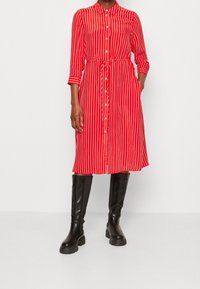 Vestito camicia a righe rosse con bottoni sul davanti, colletto e cintura in vita. Stivali neri fino al ginocchio completano l'outfit.