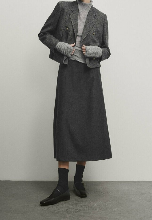 Personne portant un blazer gris court, un pull à col roulé gris avec des manches sans doigts, une longue jupe sombre, des chaussettes noires et des chaussures noires.