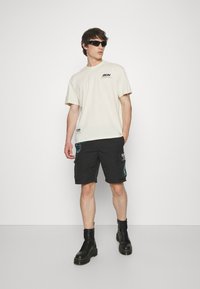 Lys beige t-skjorte i bomull med korte ermer og trykket logo. Svart cargo shorts med fargerike lapper og lommer. Svart militærstøvel.