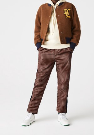 Person wearing brown varsity jacket, cream hoodie, brown plaid pants with black side panels, and white sneakers, standing with hands in pockets.