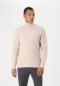 Beige gerippter Rollkragenpullover mit langen Ärmeln, featuring ein tailliertes Design und strukturiertem Stoff, kombiniert mit dunklen grauen Hosen.