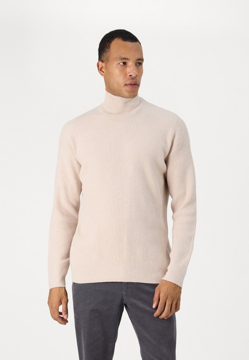 Beige gerippter Rollkragenpullover mit langen Ärmeln, featuring ein tailliertes Design und strukturiertem Stoff, kombiniert mit dunklen grauen Hosen.