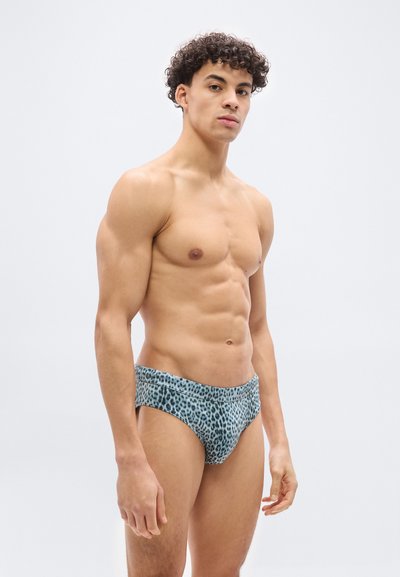 Μυώδης νεαρός με σγουρά μαλλιά, που φοράει μπλε εσώρουχα με λεοπάρ σχέδιο, στέκεται μπροστά σε απλό λευκό φόντο, κοιτάζοντας μπροστά.