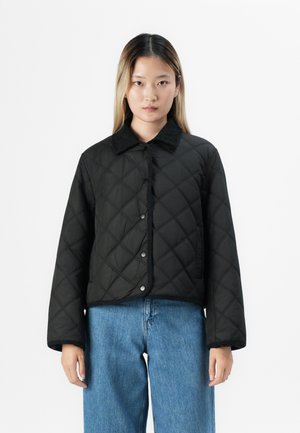 Kvinne iført en svart quiltet jakke med trykknapper og blå jeans, stående mot en ensfarget hvit bakgrunn.