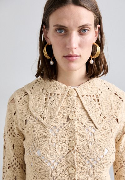 Crocheted beige cardigan with a large collar, intricate floral patterns, and three buttons down the front. Lightweight texture.