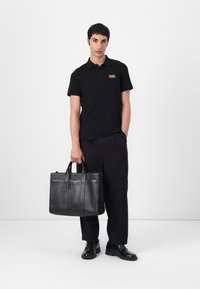 Zwarte leren tote bag met een gestructureerde vorm, dubbele handvatten en een aangehechte tag. Het model draagt een zwarte polo en broek, staand op een neutrale achtergrond.