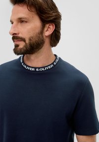 Granatowa koszulka z krótkim rękawem z ribbowanym dekoltem, z białym wzorem logo. Wykonana z miękkiej bawełny o gładkiej fakturze.