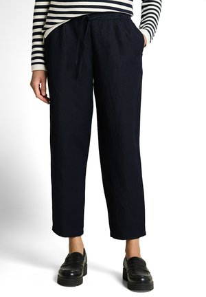 Person wearing black drawstring pants, black loafers, and a black-and-white striped long-sleeve top, standing with one hand in pocket.