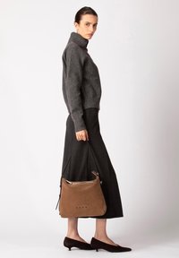 Borsa a tracolla in suede marrone con texture liscia, tracolla nera e dettagli sottili a pois sul davanti, abbinata a un maglione grigio e una gonna scura.