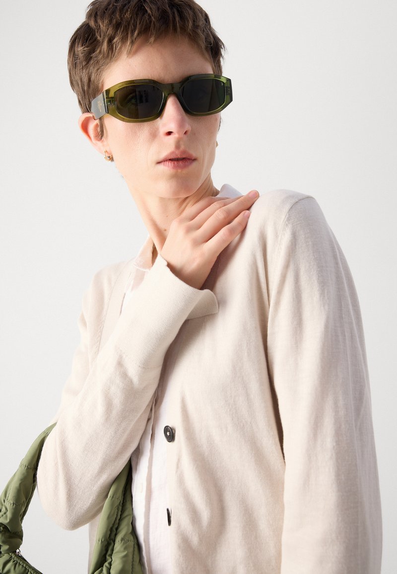 Hellbeige Strickjacke mit einer weichen Textur, kombiniert mit dunkelgrünen, eckigen Sonnenbrillen. Das Modell hält eine grüne, gepolsterte Tasche vor einem neutralen Hintergrund.