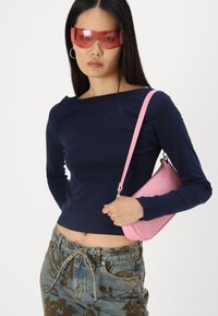 Jūros jėgos ilgarankoviai marškiniai, rožinė krokodilo tekstūros rankinė ir raštuoti mėlyni džinsai su raišteliu juosmenyje. Modelis dėvi dideles rožines saulės akinius.