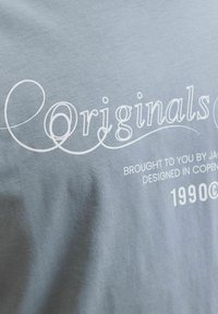 Helevalge kangas valge kirjaga, kus on sõnad "Originals", "Tootja poolt", "Disainitud Kopenhaagenis" ja "1990©" dekoratiivsetes ja lihtsates fontides.