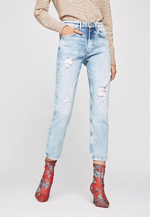 Kobieta w jasnoniebieskich przetartych dżinsach, czerwonych kwiecistych botkach na wysokim obcasie do kostek oraz beżowej bluzce w paski z długim rękawem.