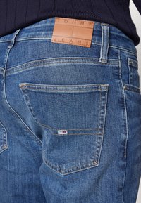 Modré džínové kalhoty s koženou nášivkou s nápisem "TOMMY JEANS" na pase a malou vlajkovou logem na zadní kapse.