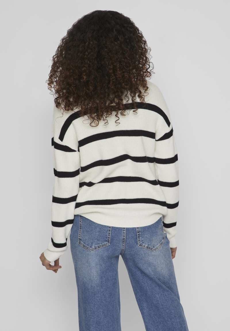 Pull crème avec des rayures horizontales noires, texture côtelée et coupe loose, associé à un jean bleu taille haute et coupe droite.