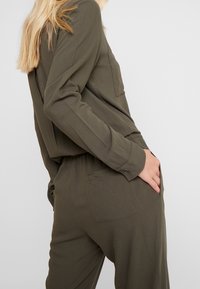 Khaki overal s uvolněným střihem, dlouhými rukávy, zapínáním na knoflíky u zápěstí a bočními kapsami. Má stahovaný pas a detail zadní kapsy.