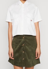Chemise blanche à manches courtes avec col, deux poches poitrine et détail logo, associée à une jupe en velours côtelé vert olive avec fermeture par boutons.