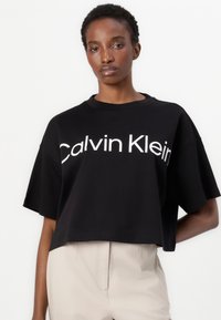 Black cropped t-shirt made from soft cotton, featuring bold white "Calvin Klein" logo across the front and relaxed short sleeves.