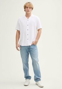 Kurzärmliges weißes Hemd mit strukturiertem Finish, kombiniert mit hellblauen Jeans und weißen Sneakers, vor einem schlichten Hintergrund stehend.