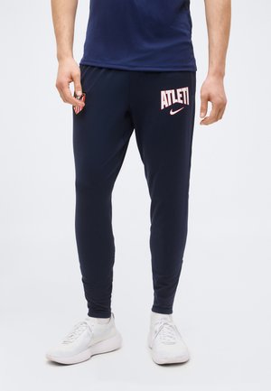 Homme portant un pantalon de sport bleu marine avec le texte « ATLETI », l'emblème du club et le logo Nike, associé à des baskets blanches, debout sur un fond blanc.