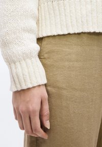 Χέρι με φυσικά νύχια στο πλάι, που φοράει ρούχο από πλεκτό κρεμ και μπεζ παντελόνι, μπροστά σε απλό φόντο.