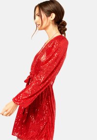 Red sequined dress featuring long sleeves, a deep V-neck, and a waist tie. The fabric has a glossy, reflective texture.