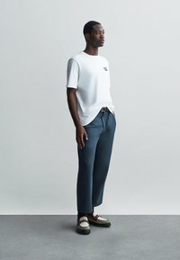 Valkoinen puuvillapaita, jossa on pieni graafinen kuvio, yhdistettynä räätälöityihin sinisiin housuihin. Jalkineissa on teksturoitu valkoisen ja ruskean yhdistelmä.