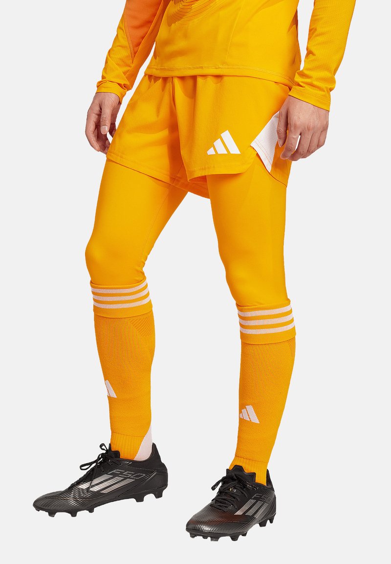 Leuchtend orangefarbensportshirt und Shorts mit weißen Streifen. Schwarze Stollenschuhe haben eine strukturierte Oberfläche. Kürzere Unterhosen sind sichtbar.