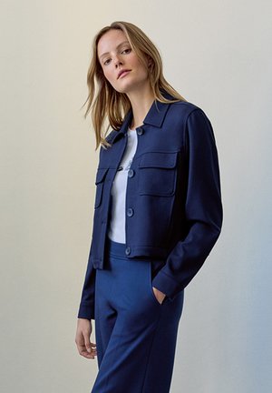 Navyblaues, tailliertes Jackett mit zwei vorderen Taschen und Knopfverschluss, kombiniert mit einem weißen Oberteil und passenden blauen Hosen. Glatte Stofftextur.