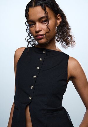 Femeie tânără cu păr creț, purtând un top negru fără mâneci cu nasturi, pozând pe un fundal simplu deschis la culoare.