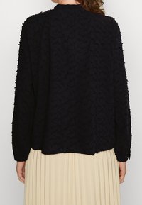 Svart texturerad blus med långa ärmar, med ett subtilt mönster. Tyget är lätt och lagrat över en neutral veckad kjol.