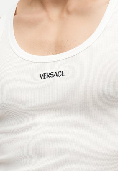 Gros plan d'une personne portant un débardeur blanc côtelé avec le logo Versace noir brodé sur la poitrine.
