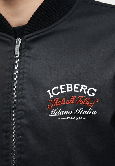 Κοντινό πλάνο μαύρου μπουφάν με φερμουάρ και κεντημένο κείμενο: ICEBERG, "That's all Folks!", Milano Italia, Established 1974.