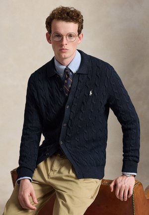 Jeune homme aux cheveux bouclés et portant des lunettes, vêtu d'un cardigan en maille torsadée bleu marine, d'une chemise bleue, d'une cravate à motifs et d'un pantalon beige, assis sur une chaise en cuir.