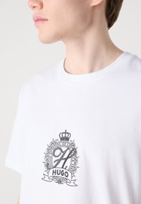 Balta kokvilnas T-kreklu ar melnu izšūtu logotipu, kurā ir kroņa un lauru dizains, teksts ir "HUGO DZĪVES SKOLA."