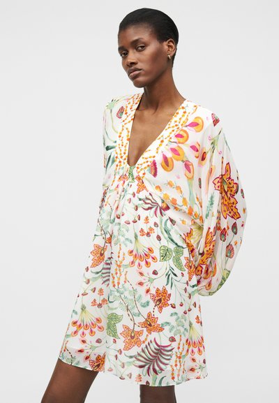 Γυναίκα που φοράει λευκό φόρεμα με πολύχρωμα λουλουδάτα μοτίβα και φαρδιά μακριά μανίκια, ποζάροντας μπροστά σε απλό φωτεινό φόντο.