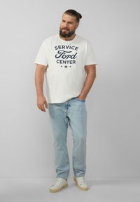 Man met een baard en een knot, in een witte "Service Ford Center" t-shirt, lichtblauwe jeans en witte sneakers, staand tegen een grijze muur.
