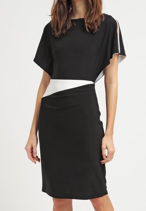 Woman wearing a black knee-length dress with a white diagonal waist panel and short flutter sleeves, standing against a plain background.