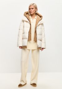 Puffjakke i hvid med en tan inderside, featuring en hætte, quilted design og front lynlås. Bæres over en kremfarvet sweater og bukser.