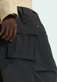 Black cargo pants feature a side pocket with textured detailing, button accents, and gathered elastic cords. Material appears lightweight.